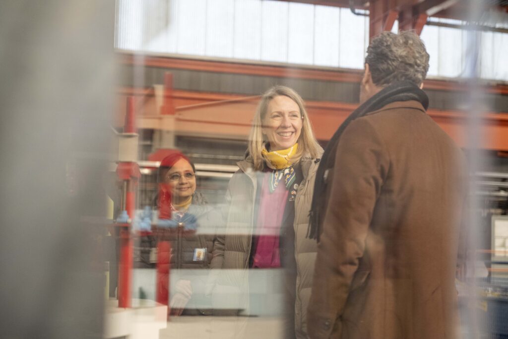 A woman with blonde hair and a scarf is seen in the reflection of a piece of acrylic, smiling at a man with his back turned to the camera. Within the display case, a scale model cyclotron particle accelerator is visible.