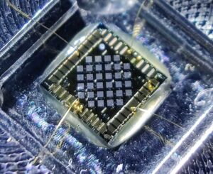 A small device resembling a computer chip, green and yellow, sits amidst an blue-tinged acrylic inlay. Small golden wires and strands connect the chip to devices out of frame.