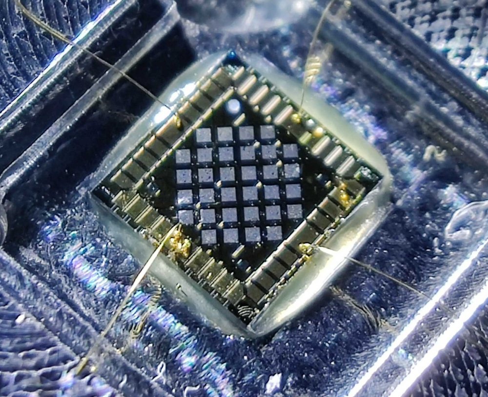 A small device resembling a computer chip, green and yellow, sits amidst an blue-tinged acrylic inlay. Small golden wires and strands connect the chip to devices out of frame.
