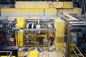 A researcher in a red jumpsuit leans over a computer next to a large apparatus made of pipes and tanks.
