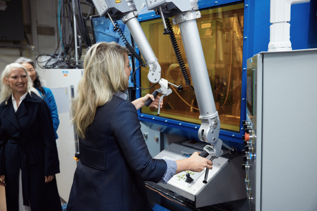 A woman with blonde hair uses telemanipulators on to move a small test sample within a giant concrete cabinet.