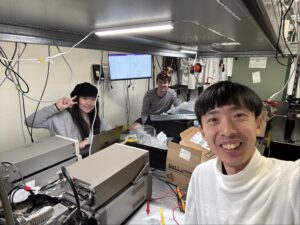 Three early-career researchers pose for a photo, a table filled with computers and wires between them.