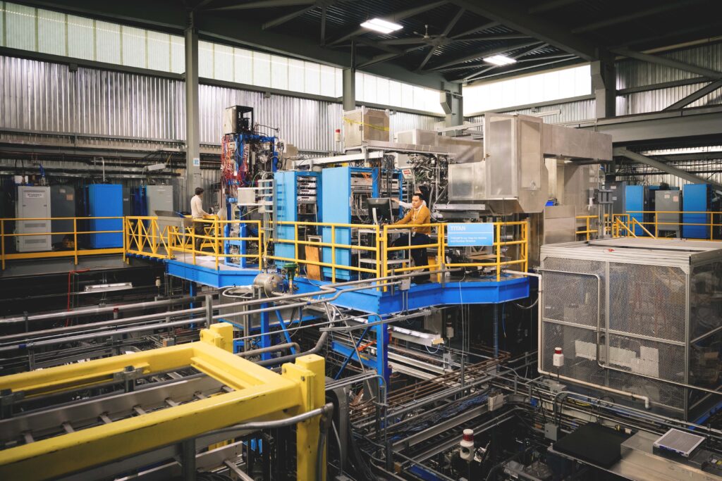 A man in a yellow sweater stands on a platform in a large science facility, looking at a laptop