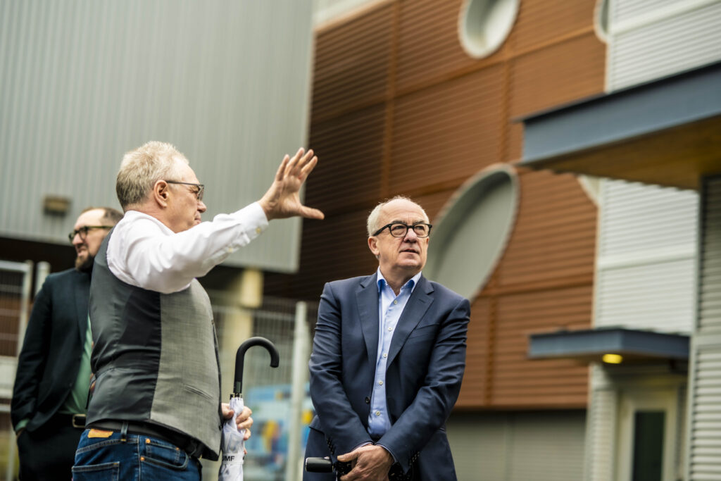 Two men stand in front of a science facility, one explaining and pointing
